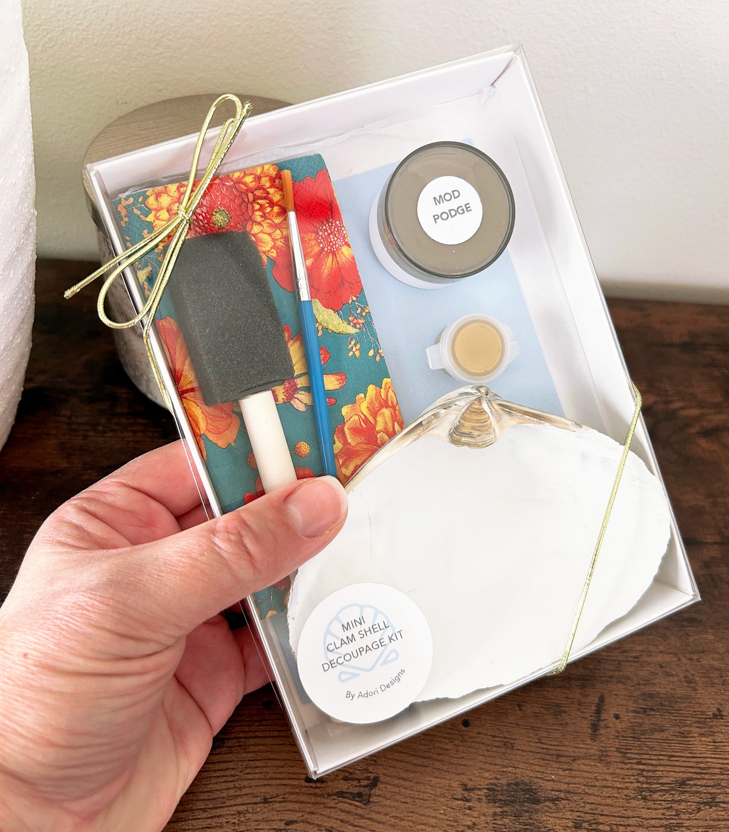 Hand holding decoupage craft kit with seashell and craft supplies on a wooden surface.