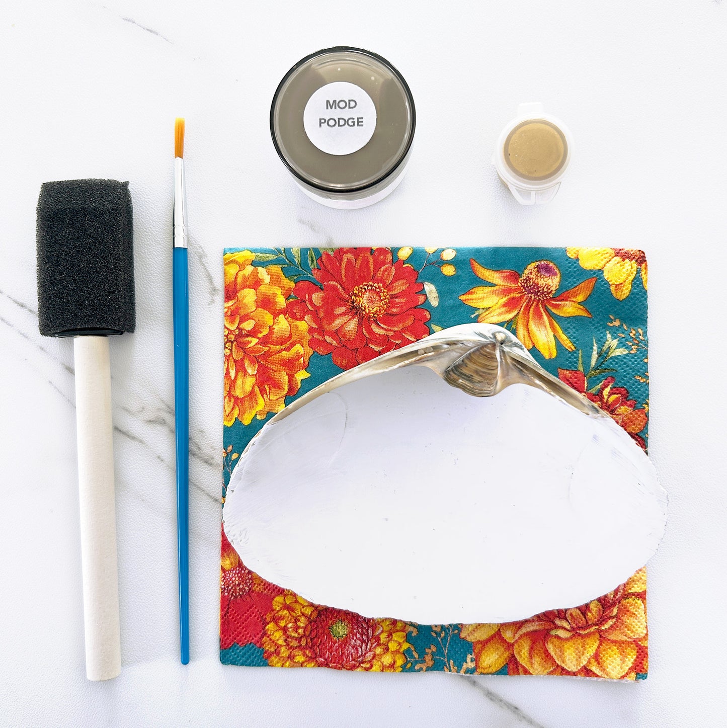 White seashell with floral design, paintbrush, and Mod Podge on a white surface
