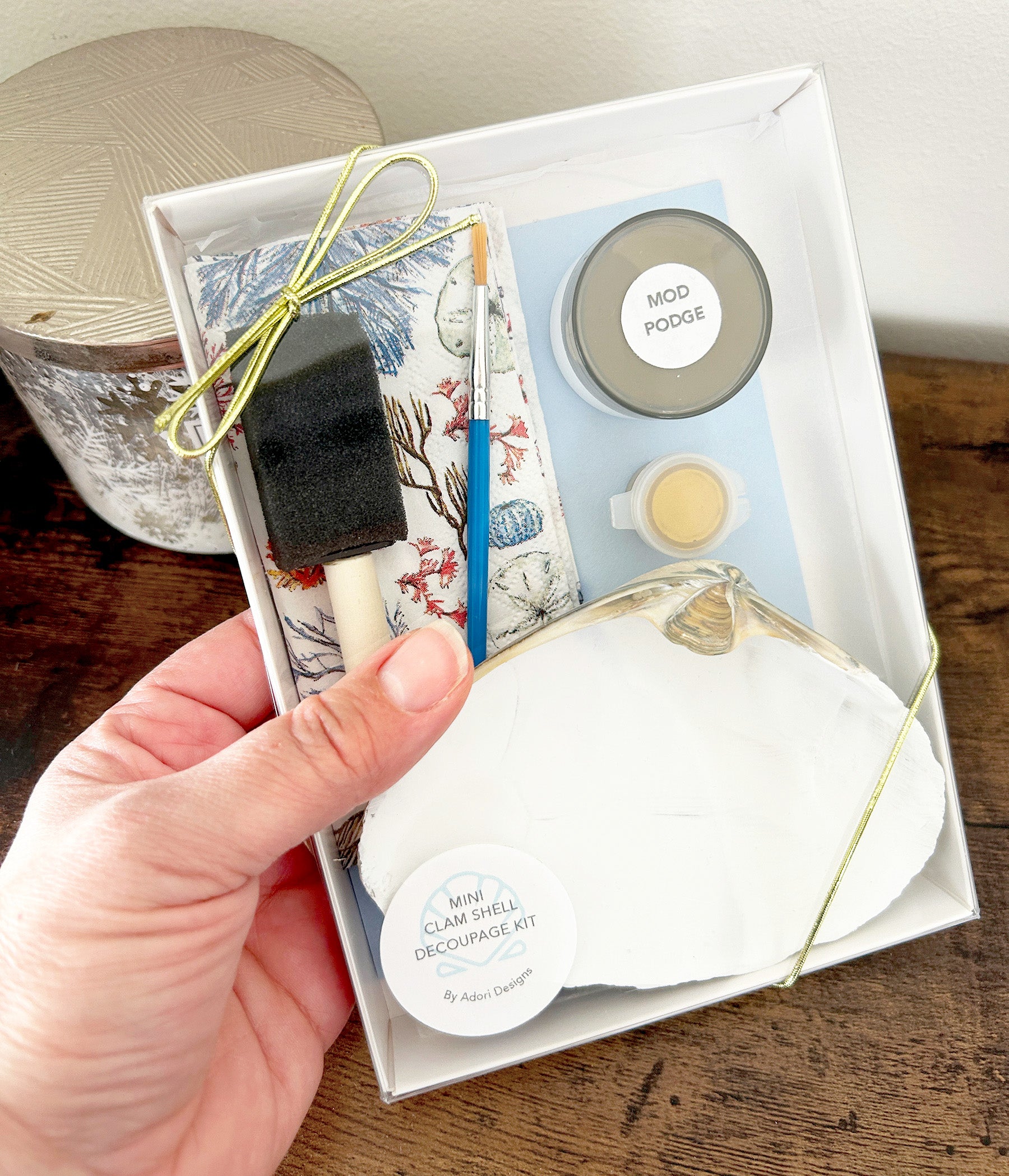 Hand holding a box of craft supplies including a sponge brush, brush, and decorative items on a wooden surface.