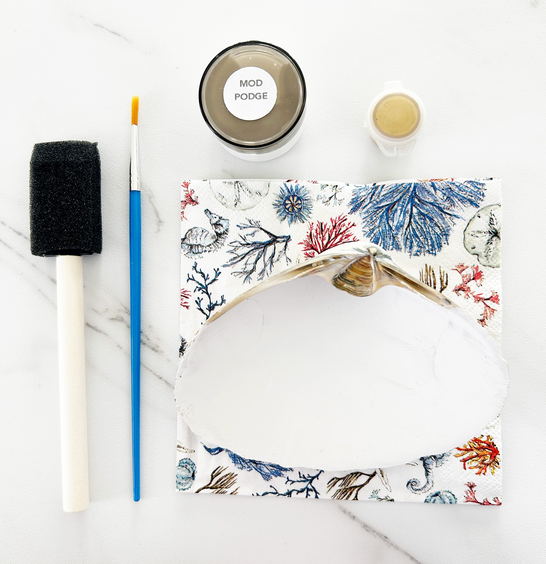White seashell with ocean-themed design, paintbrush, and Mod Podge on a marble surface