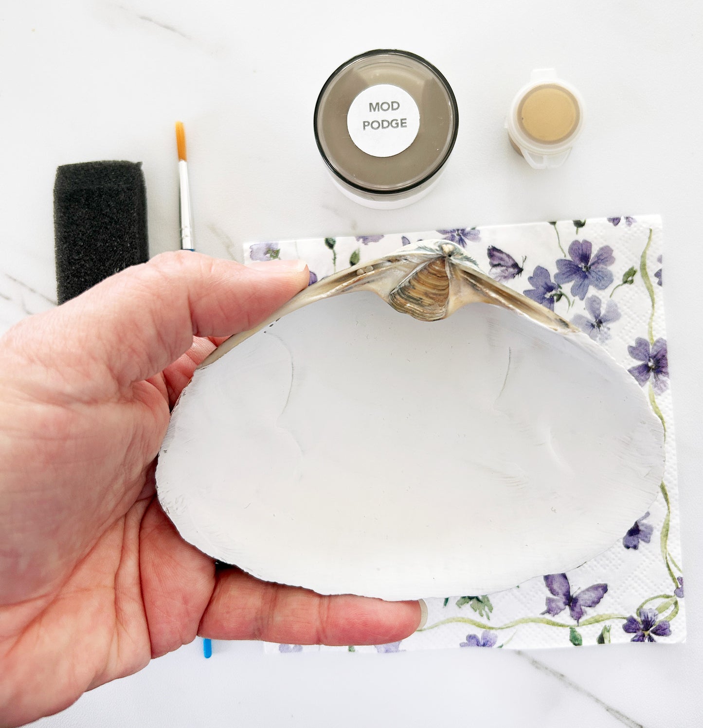 Hand holding a white seashell with floral design, surrounded by craft supplies on a white background.