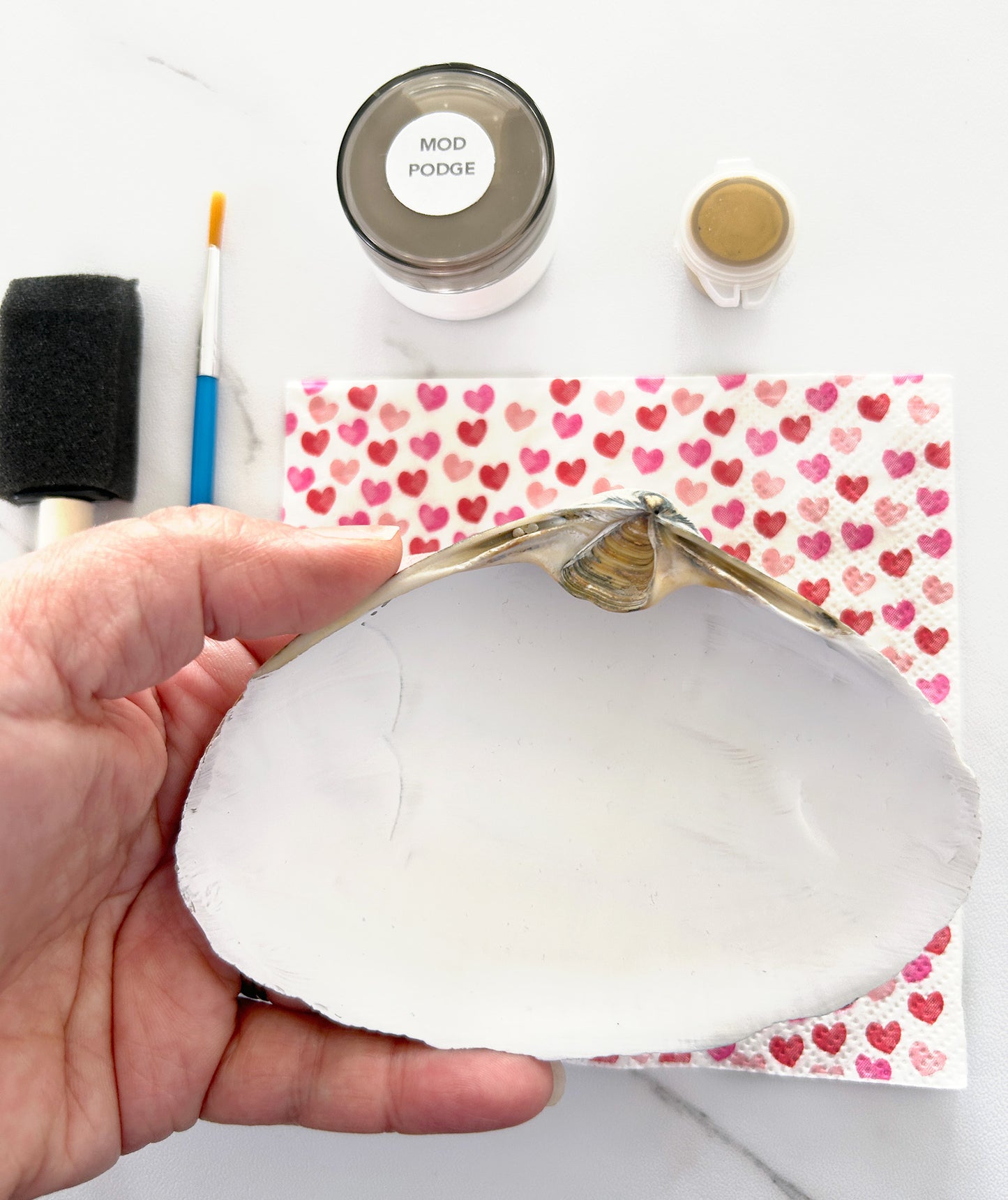 Hand holding a shell with Mod Podge and paintbrush on a heart-patterned paper