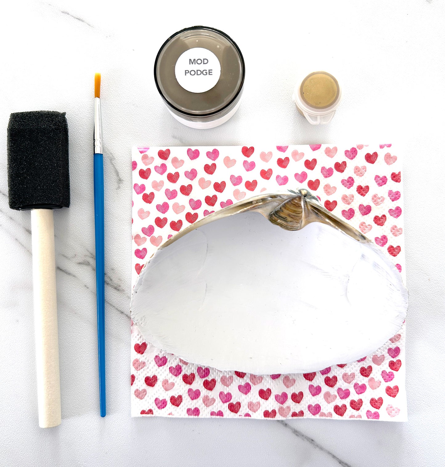 seashell with heart-patterned paper with Mod Podge and paintbrush on a marble surface